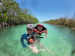 Een jongen en meisje lachen terwijl ze met zwemvesten zwemmen in helder water, omringd door mangroves en hoog gras in Mexico.
