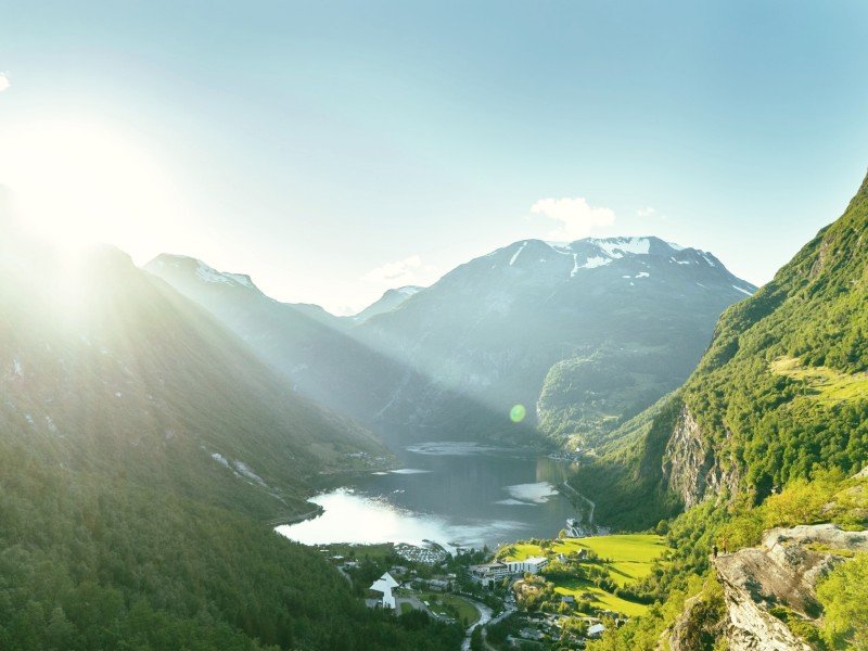 Sunlight streams over a fjord valley with green mountains and a village.