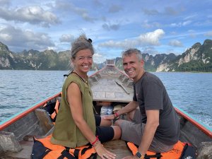 Twee reizigers zitten op een longtailboot op het water. Om hen heen rijzen steile kalksteenrotsen omhoog, begroeid met jungle. De lucht is blauw met een paar wolken, en op de houten boot liggen oranje reddingsvesten.
