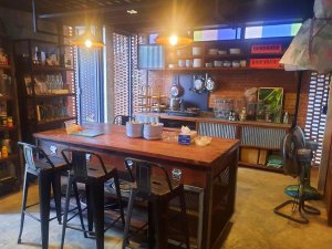 Kleurrijke, industriële keuken met een houten tafel en zwarte barkrukken in het midden. Op de achtergrond open planken met servies, een fornuis met pannen, en decoratieve elementen. Links en rechts metalen roosters voor de ramen, met lichtinval en een staande ventilator op de vloer.