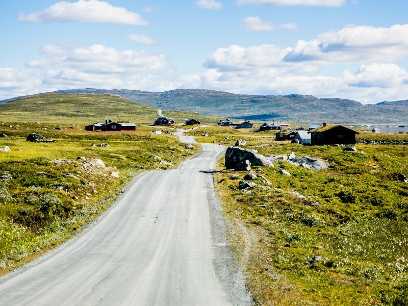 A winding dirt road leads through a green, hilly landscape dotted with cabins and distant mountains under a blue, cloudy sky.