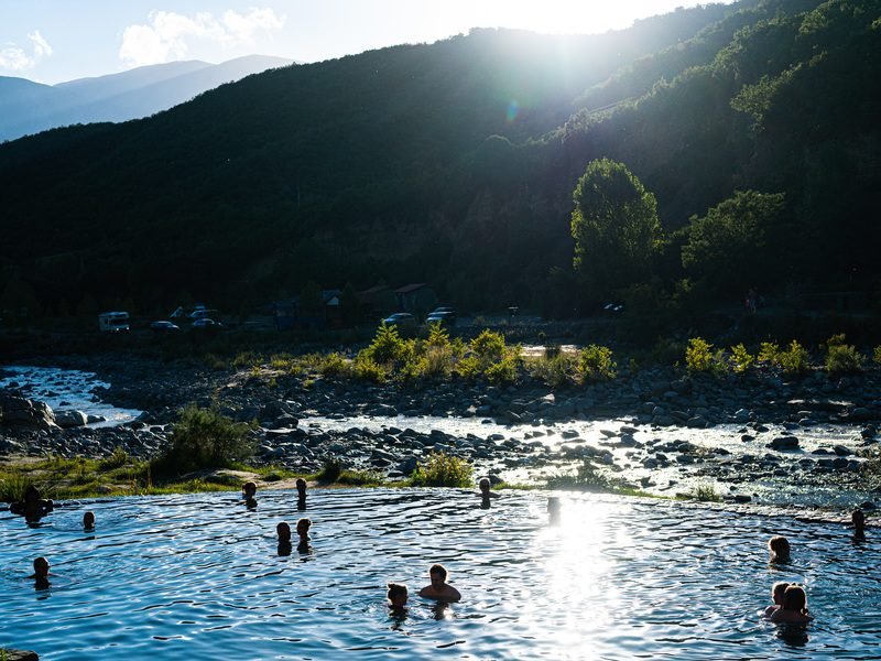 Mensen zwemmen in een natuurlijke poel in Albanië met een rivier, bergen en een camping in de verte in zonlicht