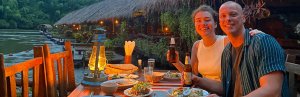 Twee reizigers genieten van een diner bij kaarslicht aan het water in een tropisch restaurant met rieten dak.
