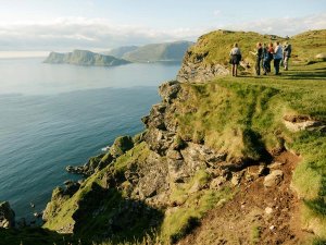 A group of people standing on a grassy clifftop overlooking the ocean and distant islands.