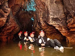 Vier mensen in wetsuits drijven op hun rug in een donkere grot en kijken omhoog naar een plafond met blauw lichtgevende glowworms in een ondergronds natuurgebied