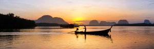Een persoon bestuurt een longtailboot, als silhouet tegen een zonsopkomst boven bergen in Thailand.