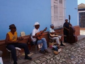 Vier muzikanten spelen snaarinstrumenten en contrabas op een kasseienstraat voor een felblauwe muur in Cuba