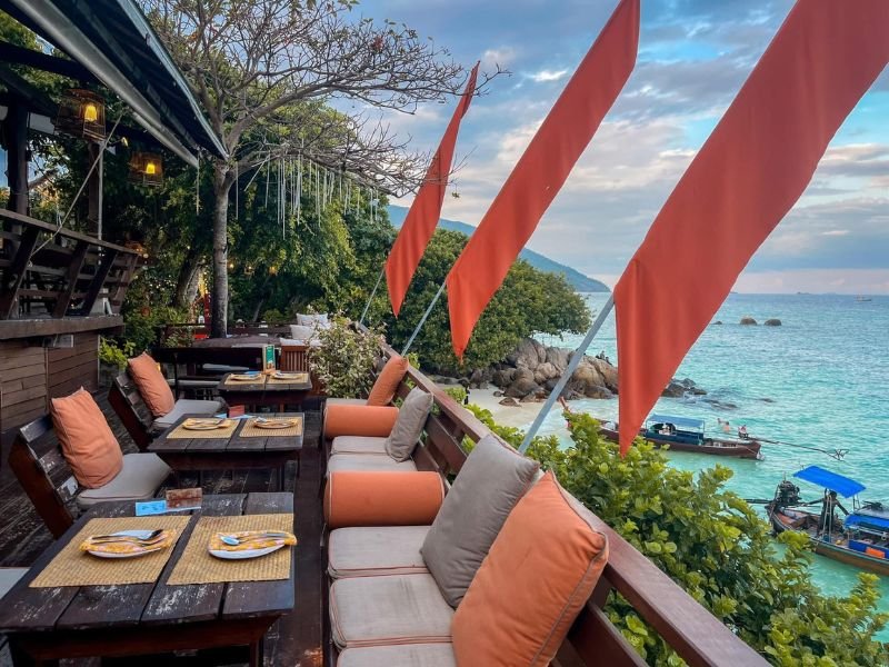 Buitenterras van een strandrestaurant met oranje en vlaggen kussens en uitzicht op turquoise zee en longtailboten.
