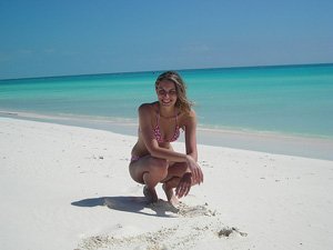 Lachende vrouw in een roze bikini hurkt op een wit zandstrand met turquoise water bij Cayo Largo in Cuba