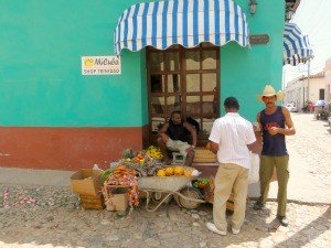 A street vendor sells produce from a wheelbarrow to customers outside a teal shop in Trinidad, Cuba.