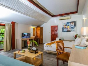 Ruime hotelkamer met een houten vloer en lichte muren. De kamer is ingericht met een groot tweepersoonsbed, houten meubels, een zitgedeelte met salontafel en een bureau met televisie. Er hangen kleurrijke kunstwerken aan de muur en op tafel staan bloemen. Een schuifdeur biedt uitzicht op tropisch groen buiten.
