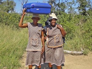 Twee lachende vrouwen in bijpassende uniformen en hoeden, waarvan één een blauwe koffer op haar hoofd balanceert op een stoffig pad in Zuid-Afrika