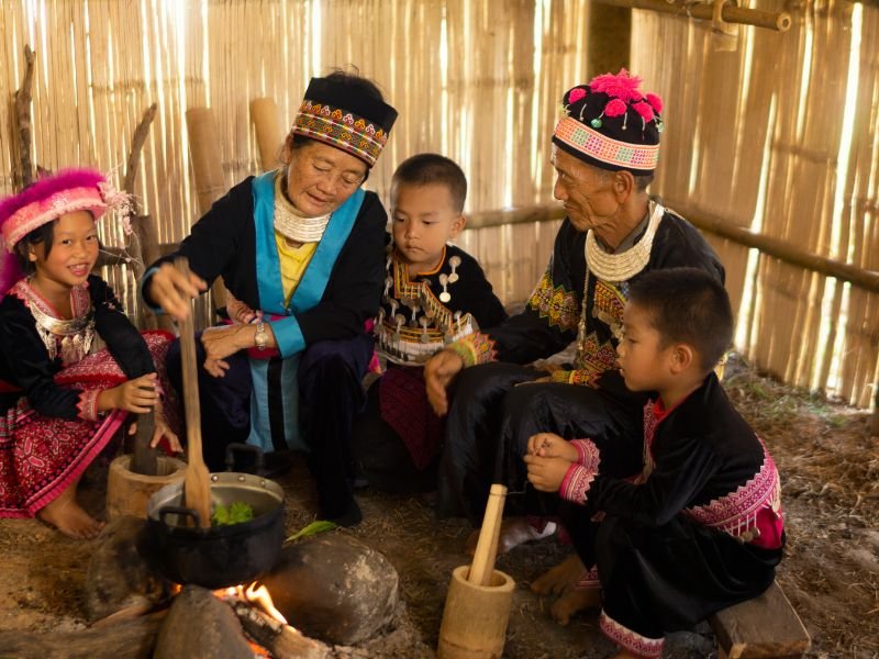 Een groep mensen in traditionele kleding zit rond een vuur in een bamboehut. Een vrouw roert in een pot; kinderen en een man zitten eromheen.