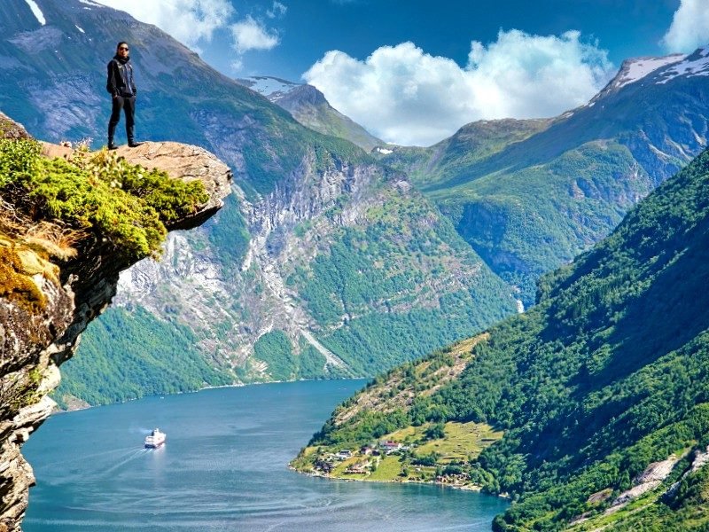 Person on a cliff overlooking a fjord with a cruise ship and green mountains.