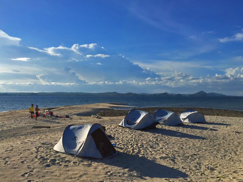 Vier uitklaptenten op een strand, met twee mensen die lopen. Zee en bergen zijn zichtbaar op de achtergrond
