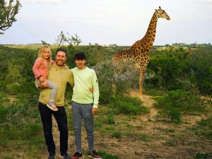 Man met een kind op de arm en een tienerjongen poseren in de savanne met een giraffe op de achtergrond in Zuid-Afrika