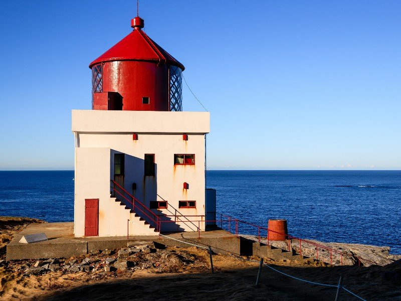 A white lighthouse with a red top stands on a rocky coast overlooking a blue sea under a clear sky.
