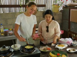 Reizigster met een lokale Thaise vrouw die naast elkaar staan te koken. De reizigster krijgt uitleg erover tijdens deze kookworkshop in Chiang Mai. Er staan op tafel allemaal bakjes met ingredienten en twee kookpitten met pannetjes erop.