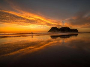 Persoon loopt over een nat strand dat een oranje en gele zonsondergang weerspiegelt met een donker eiland in de verte aan de kust