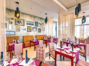 Lege restaurantzaal met ingelijste kunst, zitbanken en tafels met paarse accenten.