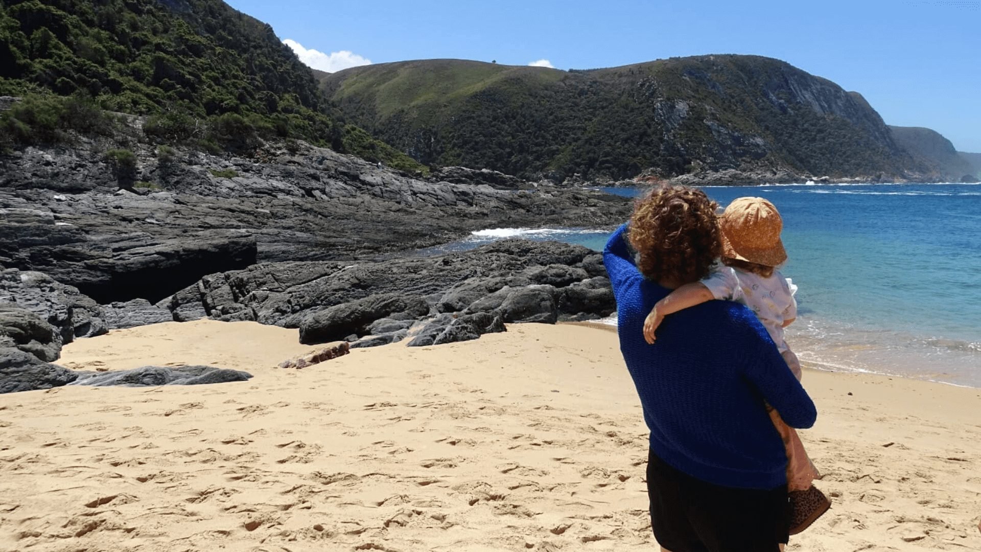 Volwassene houdt een kind vast op een zonnig strand met uitzicht op een rotsachtige kustlijn en blauwe oceaan in Zuid-Afrika