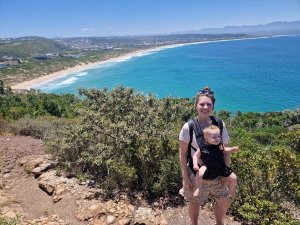 Vrouw met baby in een draagzak glimlacht op een heuvel met uitzicht over een lang zandstrand en turquoise oceaan onder een heldere blauwe lucht in Zuid-Afrika
