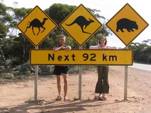 Twee mensen poseren achter een verkeersbord met waarschuwingen voor kameel, kangoeroe en wombat en de tekst Next 92 km langs de weg in de Australische outback