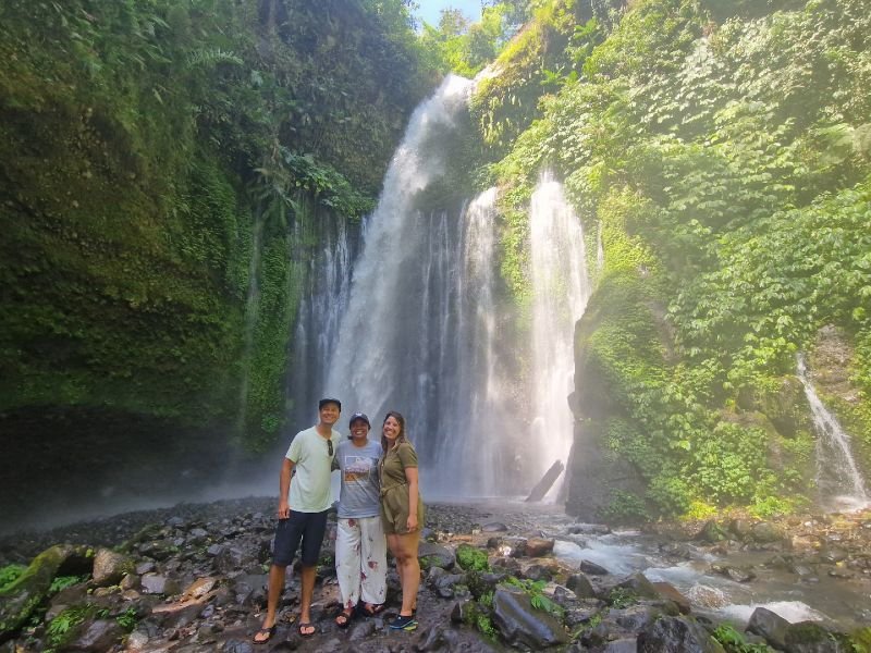 Drie mensen poseren samen voor een waterval bij Senaru op Lombok, Indonesië. Ze staan op een natte, met stenen bedekte ondergrond, omringd door mist van het vallende water. Op de achtergrond stort een hoge, krachtige waterval naar beneden tussen dichtbegroeide rotswanden met groene planten en mos. Zonlicht valt deels door het bladerdak en zorgt voor een lichte gloed in het nevelige watergordijn.