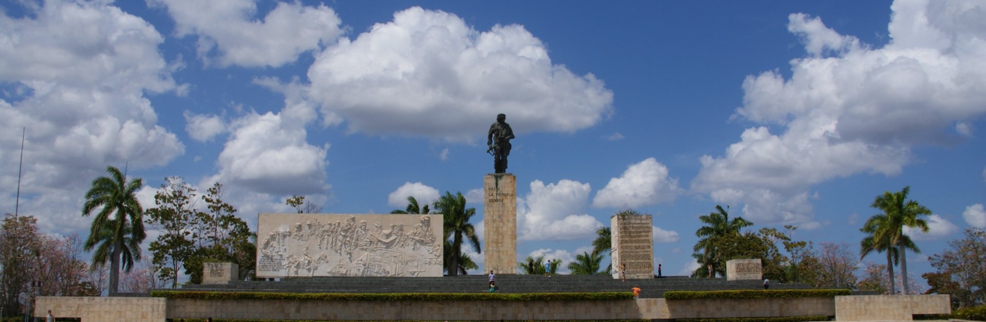 Groot standbeeld van Che Guevara met reliëf op een monument onder een blauwe lucht met wolken en palmbomen in Santa Clara in Cuba