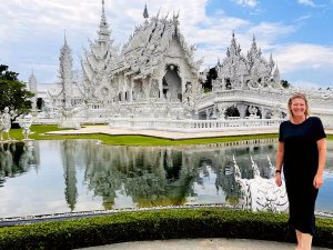 Een vrouw staat lachend voor de beroemde witte tempel in Chiang Rai, Thailand. De tempel is rijkelijk versierd met gedetailleerde, sierlijke sculpturen en wordt weerspiegeld in het water van een vijver op de voorgrond.