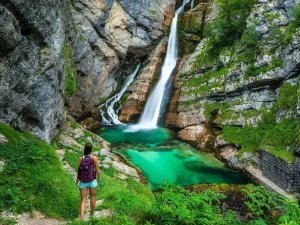 Vrouw met rugzak kijkt uit over een waterval die in een helder turquoise poel stroomt, omringd door rotsen en groen mos.