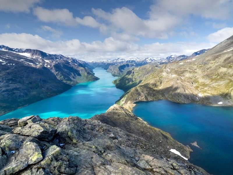 Panoramic view of a bright turquoise lake winding through rugged, snow-capped mountains, with a darker blue lake in the foreground.