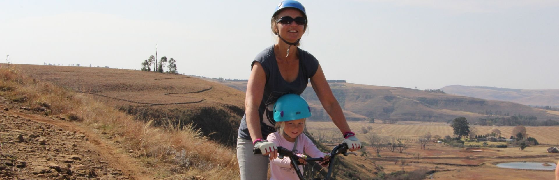 Vrouw en kind met helmen lachen terwijl ze fietsen over een zandpad door een droog, heuvelachtig landschap in Zuid-Afrika