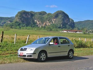 Vrouw lacht vanuit een zilveren auto langs de weg met groene velden en kenmerkende ronde heuvels op de achtergrond in Cuba