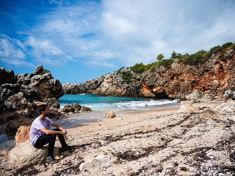 Man op een rots op een rotsachtig strand in Albanië die uitkijkt op een natuurlijke rotsboog boven een helderblauwe baai