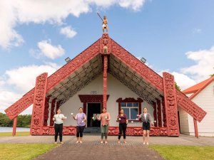 Vijf vrouwen houden poi vast voor een traditioneel gesneden Māori wharenui met een beeld op het dak in een culturele setting in Nieuw-Zeeland