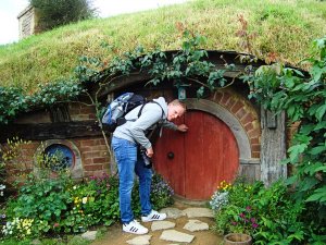 Man met rugzak en camera klopt op de ronde rode deur van een hobbitwoning in een grasheuvel in Nieuw-Zeeland