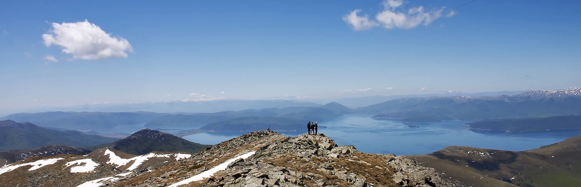 Panoramisch uitzicht vanaf een rotsachtige berg met sneeuwplekken, waar twee mensen op een top staan met uitzicht op een groot meer en bergen in de verte in Nationaal Park Pelister in Noord-Macedonië