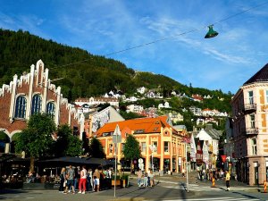 A lively city street with historic, colorful buildings and people, set against a forested mountain dotted with houses under a blue sky.