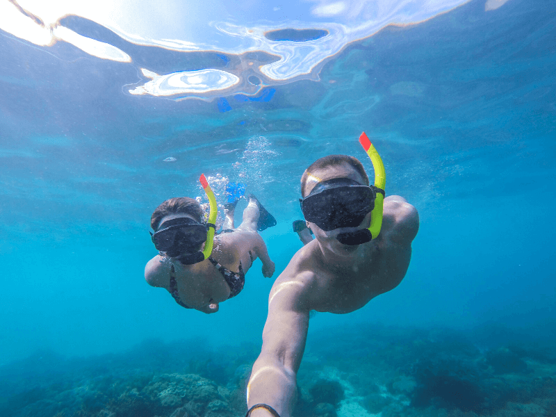 Twee snorkelaars zwemmen zij aan zij in helderblauw tropisch water. Ze dragen duikbrillen met snorkels en maken een selfie onder water. Op de achtergrond zie je de zee langzaam vervagen in de diepte, met koralen zichtbaar op de bodem.