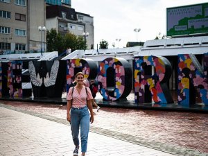 Lachende vrouw in een roze top en jeans loopt langs het kleurrijke NEWBORN-monument met gezichten en witte vleugels in Pristina in Kosovo