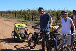 Twee volwassenen met fietsen en een kindertrailer staan op een zandpad naast een wijngaard in Zuid-Afrika