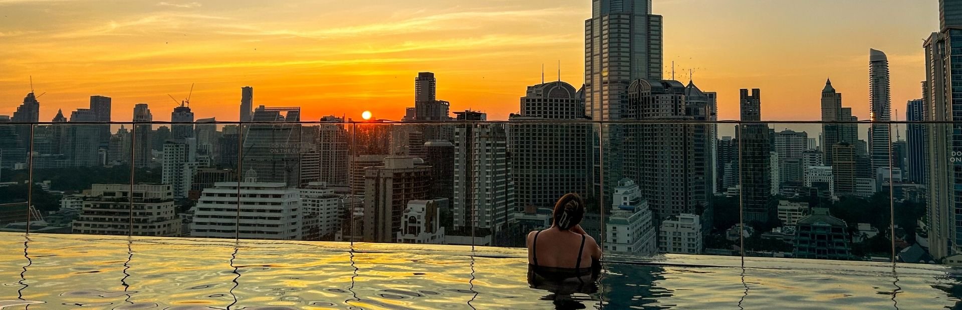 Infinity pool op een dakterras met uitzicht op de skyline van Bangkok. Vrouwelijke reiziger dobbert aan de rand en kijkt uit over de zonsondergang.