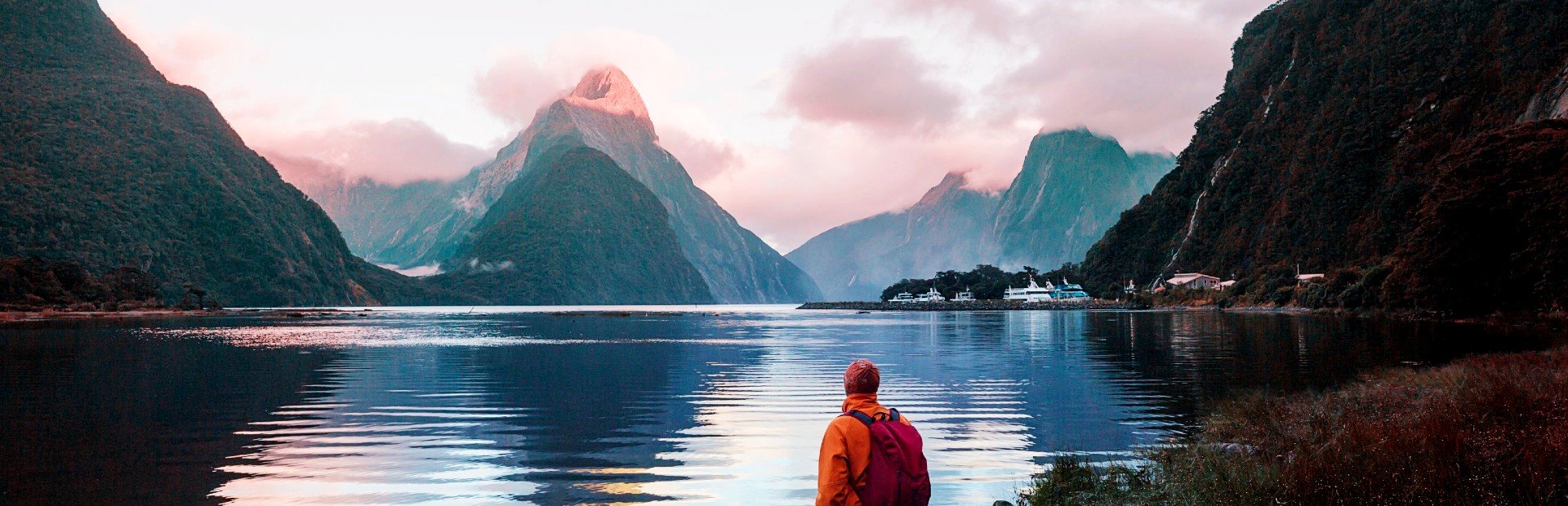 Persoon kijkt uit over een indrukwekkende fjord omringd door bergen met een bergtop die roze oplicht bij zonsopgang in een bergachtig natuurgebied