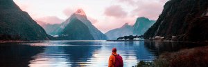 Persoon kijkt uit over een indrukwekkende fjord omringd door bergen met een bergtop die roze oplicht bij zonsopgang in een bergachtig natuurgebied