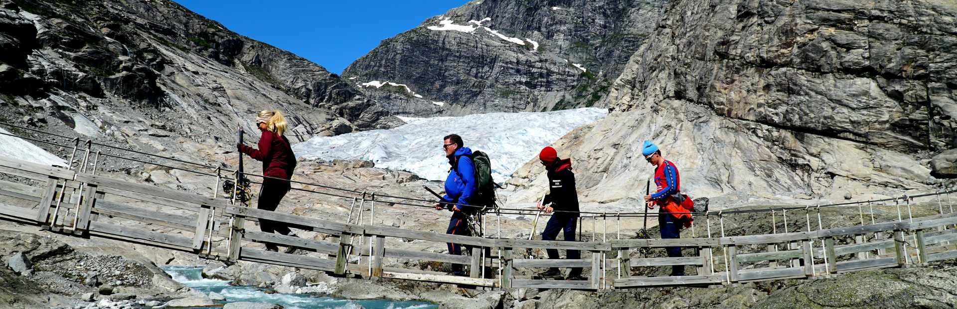 Four hikers crossing a wooden bridge over a glacial river, with a large glacier and rocky mountains in the background.