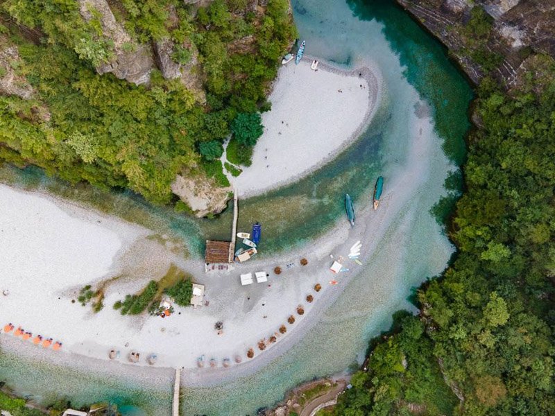 Luchtfoto in Albanië van een kronkelende turquoise rivier door groene bossen met een zandstrand, boten en kleine gebouwen