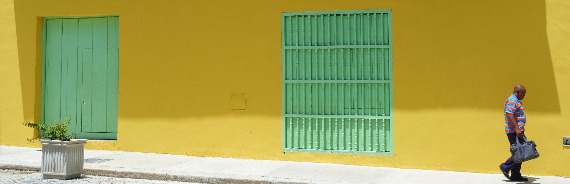 Man loopt langs een felgele muur met een groene deur en een groen raam met tralies in Cuba