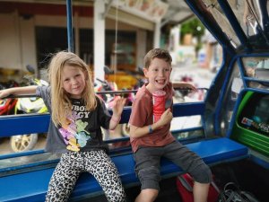 Twee jonge kinderen zitten vrolijk naast elkaar op een blauwe bank achterin een Thaise tuktuk of songthaew (openbaar vervoersbusje). Het meisje links draagt een zwart T-shirt met kleurrijke cartoonfiguren en een luipaardprint broek, en leunt nonchalant met haar arm op de rugleuning. De jongen rechts draagt een rood T-shirt en een grijze korte broek, lacht breed en houdt een rood Pringles-busje vast. Op de achtergrond zijn geparkeerde scooters en een winkelpand te zien, wat een typisch straatbeeld in Thailand geeft. De sfeer is speels en ontspannen, passend bij een familiereis.