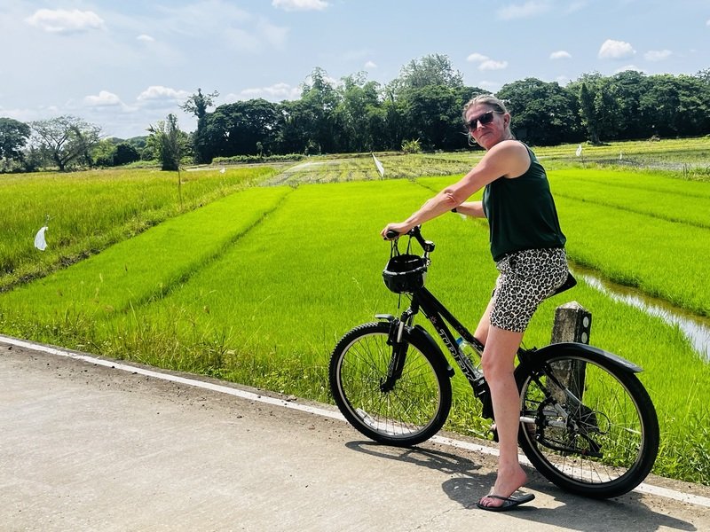 Een vrouw zit op een zwarte fiets op een betonnen weg, met felgroene rijstvelden op de achtergrond. Ze draagt een donkere top, een korte broek met panterprint en een zonnebril. Het is zonnig weer.